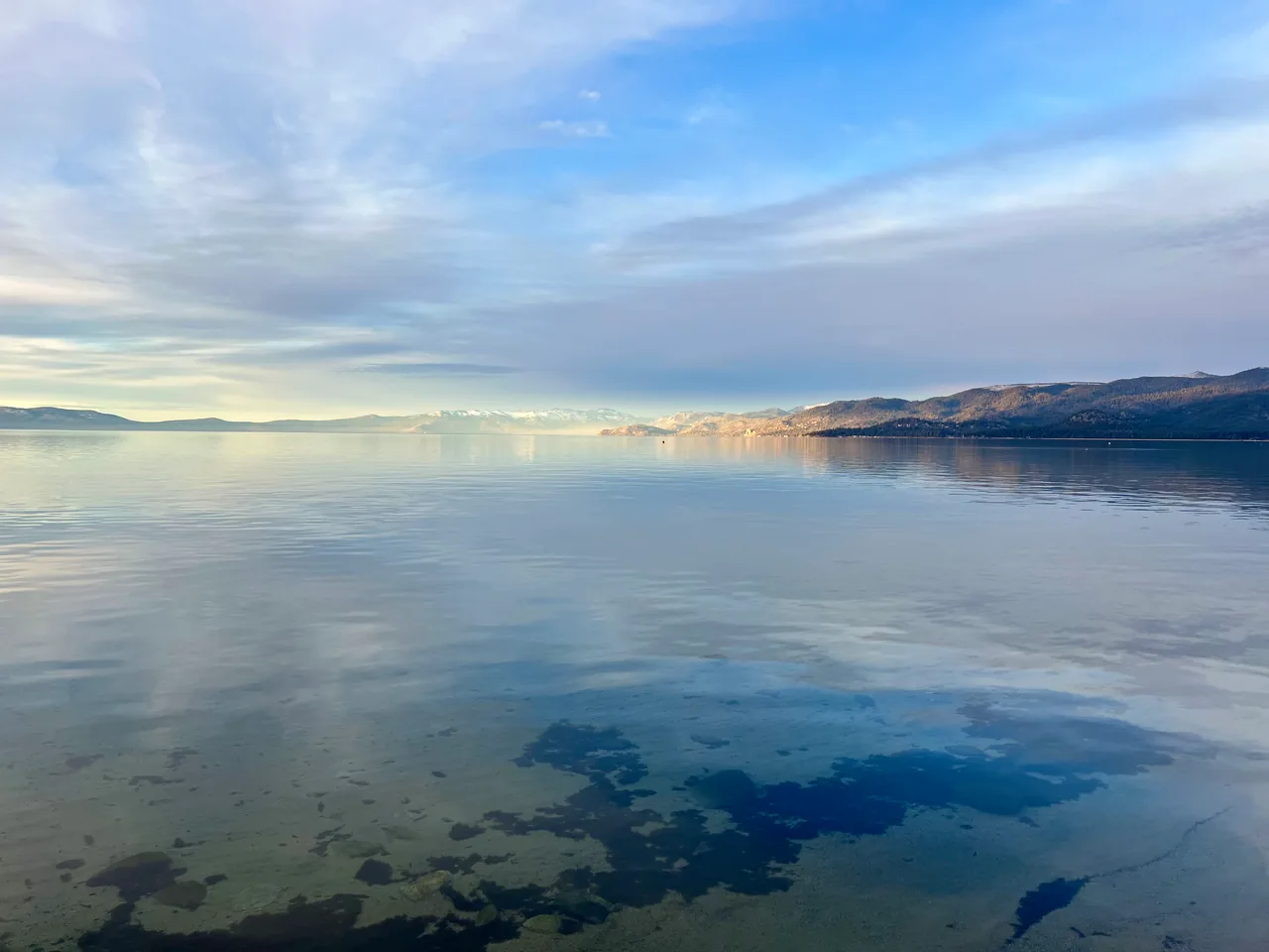 Clear shallows opening onto a calm lake beneath pale morning clouds