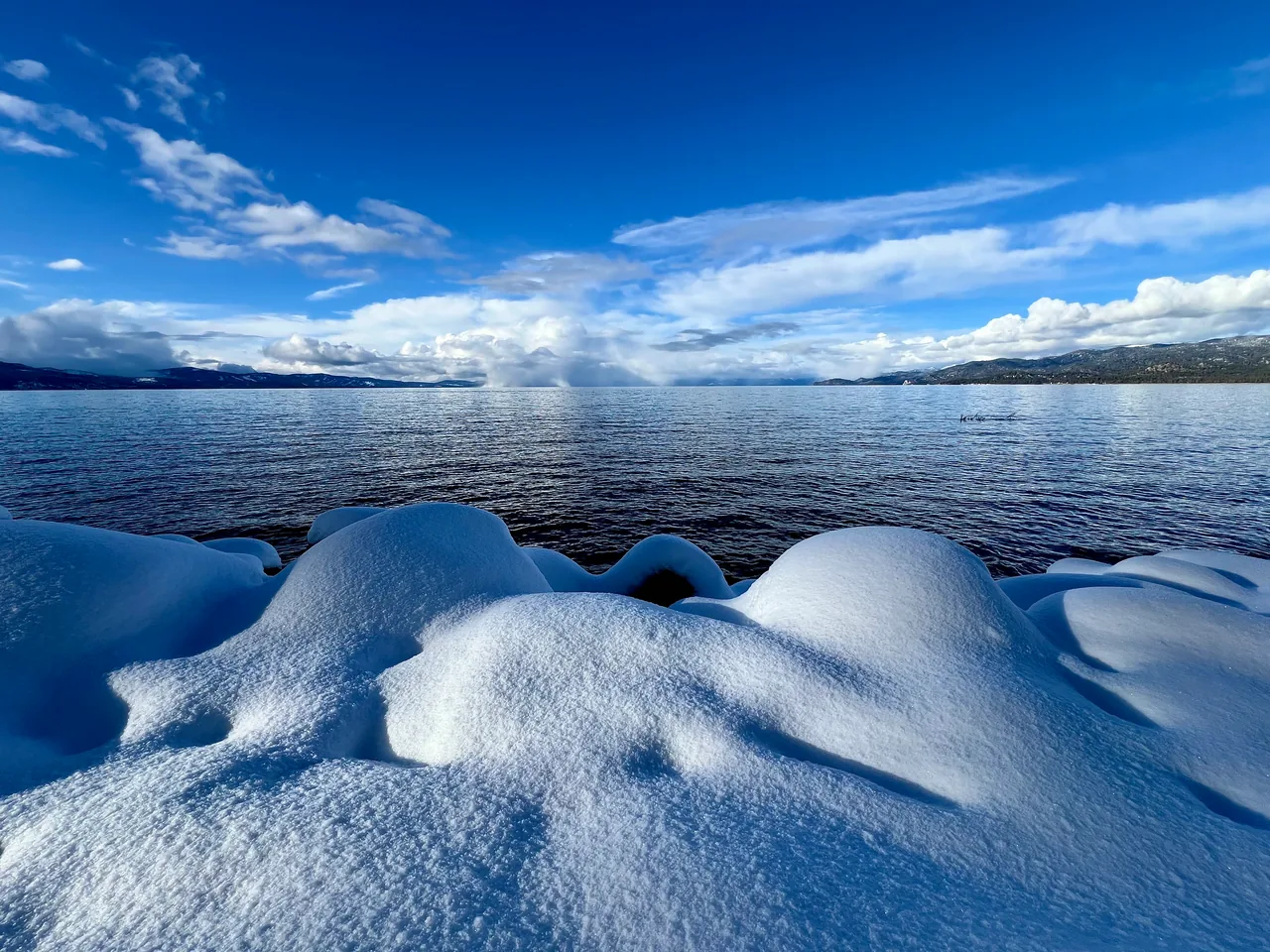 Rounded snowbanks on a lakeshore beneath a blue sky with clouds