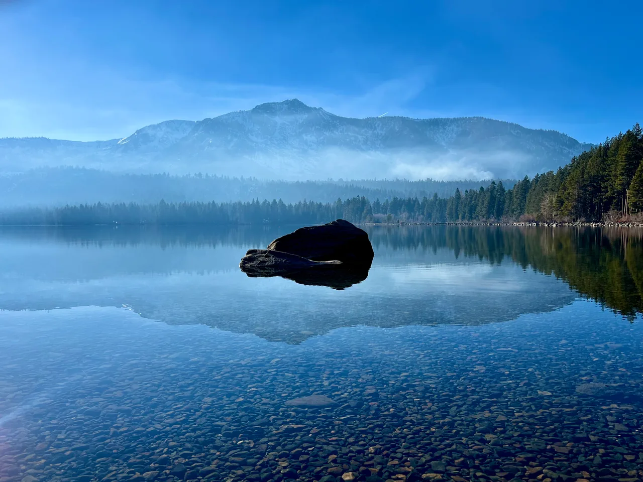 A dark boulder reflected in glassy water beneath misty mountains